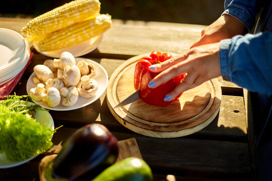 Unrecognizable woman cutting fresh pepper vegetables on wooden board during weekend barbecue in yard, outdoor, prepare for grilling, summer family picnic, food on the nature.