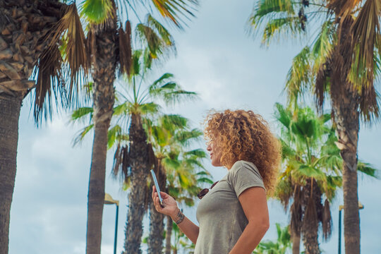 Side portrait of tourist using mobile phone in outdoor with palms street road in backgorund. One female people messaging happy. Concept of tourism and summer tropical holiday vacation leisure