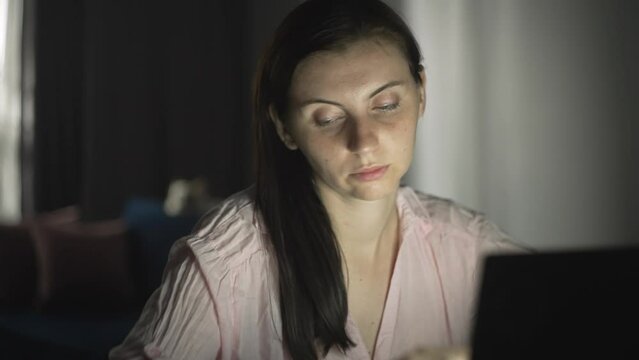 The girl works at the computer at home at night. Close-up of the face of a brunette at work. Light from a laptop. The woman is tired of working at the computer, evening work. 4k