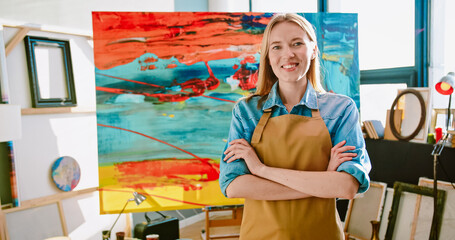 Portrait of cheerful young Caucasian beautiful talented woman artist in apron standing in studio workshop looking at camera and smiling in good mood, female painter with happy face