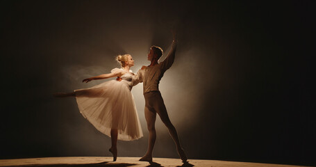 Professional duet of ballet dancers performing on stage isolated on smoked black background. Young...