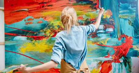Fotobehang Schilderij Close up of Caucasian cheerful happy young female artist in apron working in art studio painting using paint brush on big canvas making emotional colorful abstract masterpiece. Modern art concept  © ihorvsn