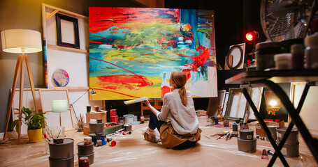 Back view of talented concentrated Caucasian woman professional artist sitting on floor in studio in evening painting drawing on big canvas, creativity work, contemporary creative painter