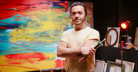 Portrait of handsome cheerful young Caucasian talented man painter holding in hands many paint brushes just finished contemporary colorful painting on big canvas in studio in evening. Artist concept