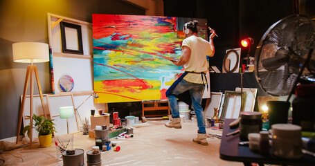 Joyful Caucasian young talented male artist listening to favorite song and dancing while drawing oil painting on big canvas in cozy studio. Creative man painter creating abstract artwork in workshop - obrazy, fototapety, plakaty
