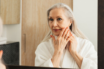 Senior woman with long grey hair touching her cheek with fingers in bathroom - obrazy, fototapety, plakaty