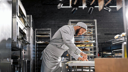 Handsome Caucasian man in white uniform cutting dough and shaping cookies in bakery. Baking concept. Indoor. Male good-looking baker producing bread or pastry in cozy comfortable kitchen.