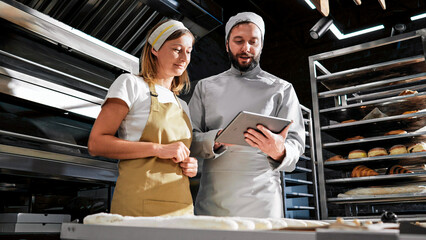Caucasian handsome man baker in uniform showing something on tablet device to woman colleague. Bakers talking and discussing work with computer in hands. Bakery concept. Workday at bakeshop.