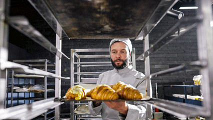 Caucasian handsome man in hat and uniform working in bakehouse and putting croissants on shelf. Male baker making fresh pastries in kitchen of bakeshop in morning. Confectionery producing concept.