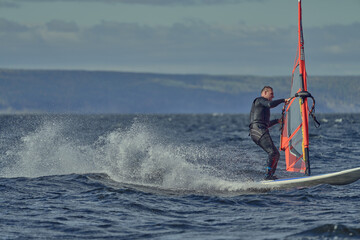 Obraz premium A mature man, an experienced windsurfer, rushes along the surface of the water, splashes scatter to the sides, a sunny autumn day.