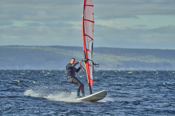 A mature man, an experienced windsurfer, rushes along the surface of the water, splashes scatter to the sides, a sunny autumn day.