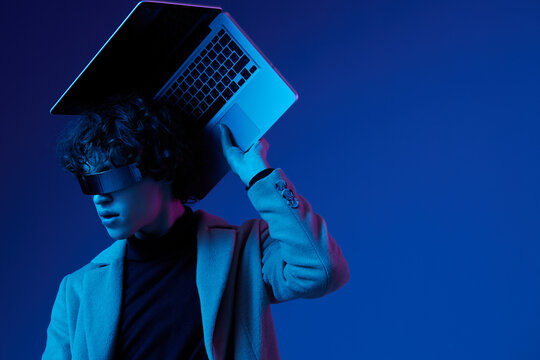 Portrait of a stylish male hacker with a laptop and futuristic glasses in blue light, cyber security, technology and education, neon color, freelancer online work, trend color