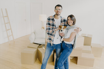 Fototapeta na wymiar Happy woman and man tenants or renters of flat pose in own house, cuddle and pose with little dog, have glad expressions, start living in new bought apartment. Family and relocation concept.
