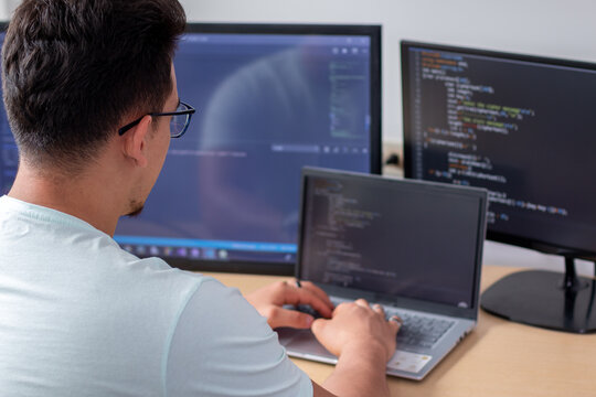 A Young Programmer Wearing Glasses Working With Three Monitors, Writing Programming Code.