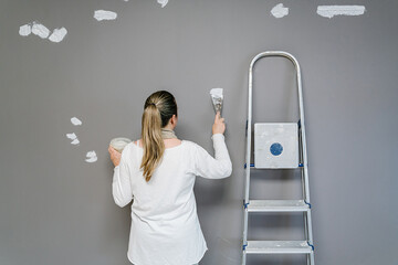 Backwards Blonde woman plastering with a spatula a wall after being painted with a grey painting. The painter is next to her ladder while covering small holes on the wall. Horizont