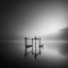 ultra wide angle long exposure black and white photograph of a lake without waves with pillars sticking out of the water inside with fog and a cloudless sky