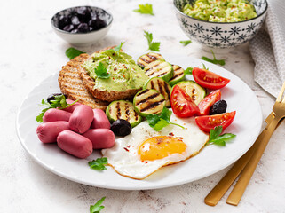 Fried egg with mini sausage, tomatoes, grilled zucchini, black olives and toasts with guacamole. English Breakfast. Brunch.
