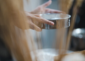 Young woman standing in kitchen sifting flour for homemade bakery or bread baking ingredient. Asian female chef holding sieve sifts flour preparing for egg mixing in process of making pastry or cake.
