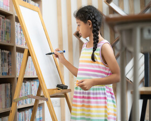 Young girl student standing in classroom writing on a whiteboard. Portrait of happy Asian schoolgirl kid holding marker pen write on blank whiteboard. Child and elementary school education lifestyle.