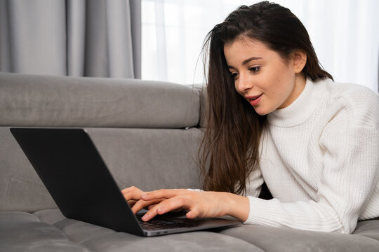 Happy young woman lying on sofa chatting on social network or shopping online using laptop at home with modern interior