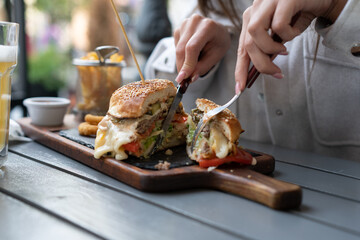 A girl is slicing her delicious burger with lots of toppings using a knife and fork.