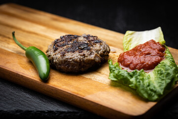 Bulgarian grilled ground meat patty on wooden board next to green chili pepper and lettuce leaf with traditional homemade dipping spread lutenica
