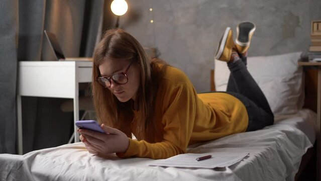 teenager girl student in glasses lies on a bed in a college dormitory holding a smartphone in her hands and communicates with friends online in social networks, writes and reads a message.