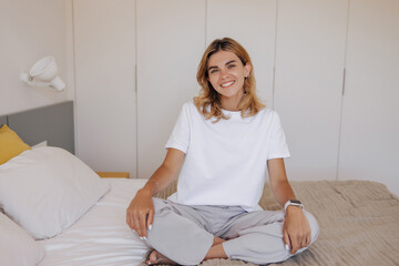 Happy young caucasian woman sitting on bed and wide smiling at camera indoor. Blonde hair girl with...