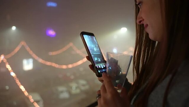 Trendy cellphone in hands of pretty smiling woman standing on balcony against amazing illuminated streets background, woman taking pictures or recording video of festive street illumination