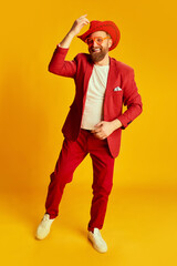 Fashionable man, hipster wearing bright red costume and cowboy hat posing over yellow background. Concept of style , fashion, happy mood, positive emotions