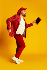 Dance. Fashionable man, hipster wearing bright red costume and cowboy hat posing over yellow background. Concept of style , fashion, happy mood, positive emotions