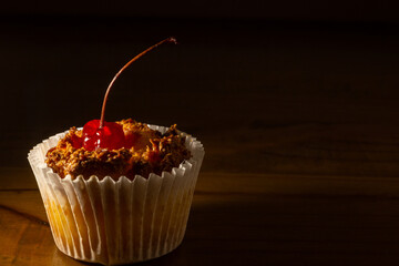 Vista lateral de um bombocado, doce típico brasileiro, com uma cereja em cima, sobre uma mesa de madeira e com luz direcionada ao produto.