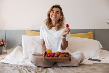 Happy young caucasian woman sitting on bed with strawberries and breakfast tray indoor. Girl wears pajamas looking at camera wide smile. Concept healthy morning