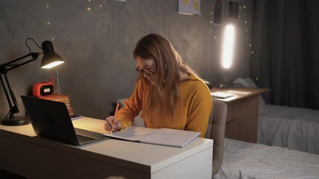 girl teenager student sits at a table in a college dormitory looking for information online using a laptop and notes in a notebook.