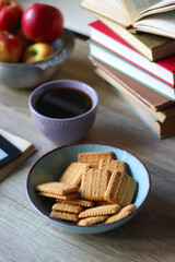 Stack of books, e-reader, reading glasses, bowl of biscuits, apples and cup of tea on the table. Bookshelf in the background. Selective focus.