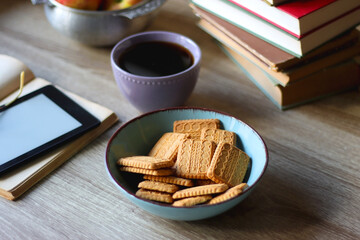 Stack of books, e-reader, reading glasses, bowl of biscuits, apples and cup of tea on the table. Bookshelf in the background. Selective focus.