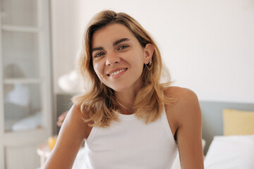 Close-up cheerful young caucasian girl wear white t-shirt looking at camera with smile indoor. Woman blonde hair posing on bed in bedroom. Concept portrait, beauty, lifestyle.