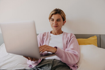 Calm young caucasian female holds laptop sitting on bed in bedroom morning. Blonde woman texting massages on laptop at home. Concept self-employment.