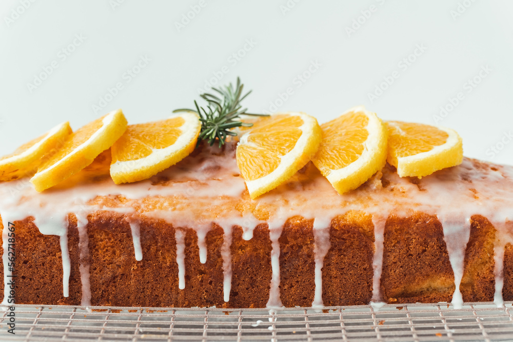 Wall mural Loaf of orange bread covered with a confectionery glaze with lemon juice and decorated with orange slices. Chiffon cake on a pastry grill next to cup of tea. White background - Wall murals