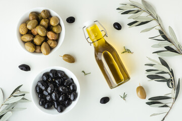 Green and black olives in white bowls next to a bottle with olive oil and leaves on a white background. .Bottle of cold pressed oil. Traditional Greek and Italian food. Flat lay.