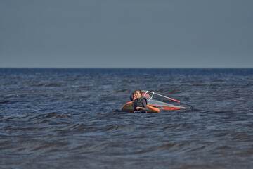 Mature woman windsurfer novice walks through shallow water and pulls a sailboard behind her on a sunny autumn day.