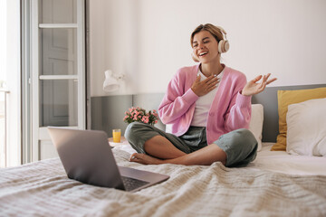 Friendly young caucasian woman with wireless headphones, using her laptop. Blonde hair girl sitting on bed against window. Concept modern technologies.