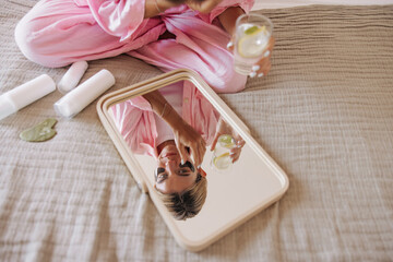 Top view young caucasian girl reflected in mirror sitting on bed and applying face cream. Blonde hair woman holds glass water with slice of lemon. Concept beauty procedures at home.