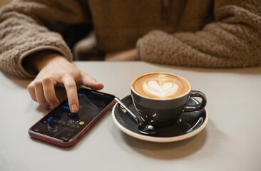 Girl in fluffy sweater writes on smartphone kissing emoji message next to cup of coffee with heart shaped latte art foam. Closeup cup of coffee with cream in coffee shop. St. Valentine's Day. Love