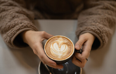 Young woman in a fluffy sweater holds in hands a cup of coffee with heart shaped latte art foam. Close up cup of coffee with cream in coffee shop. Valentine's Day. Love. Date