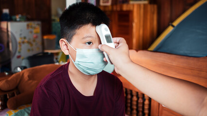 A mother's hand using an infrared thermometer measures the body temperature of an Asian son wearing a mask because he is ill.