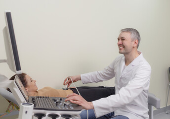 A male doctor moving an ultrasound sensor on a pregnant woman's stomach while looking at a screen in a hospital.