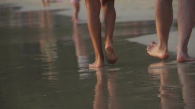 Close up detail bottom behind pov view of female and male person barefoot legs heel walking by scenic sandy ocean or sea beach at warm golden sunrise morning. Wild nature travel and vacation concept