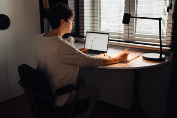 Senior woman working on laptop computer and writing down notes while sitting at table