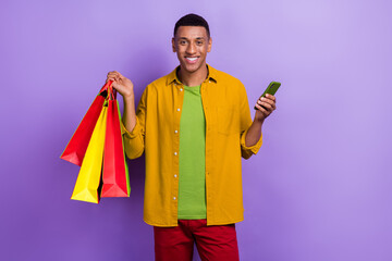 Portrait of good mood handsome guy wear yellow clothes holding shopping bags smartphone online order isolated on violet color background